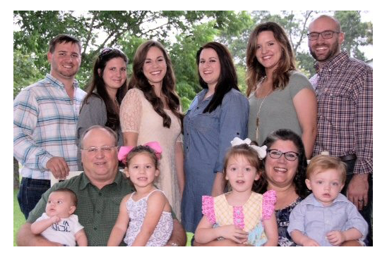 Photo of James’ family: (standing left to right) son, Ethan, daughter-in-law, Jaci, daughters, Chrissy and Mel, daughter-in-law, Courtney and son, Billy; (sitting left to right) James holding grandchildren, Dawson and Cheyenne, and James' wife Ellen holding grandchildren, Everly and Wilder.